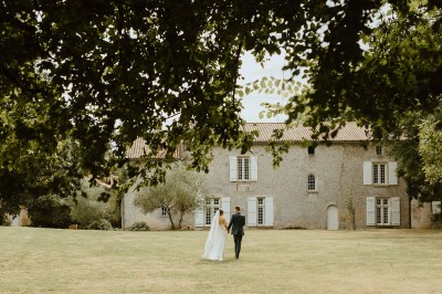 Miniature Location salle L'Orbrie (Vendée) - Le Logis des Arçonnières #79 Un couple marche dans un jardin verdoyant en direction d'une grande maison en pierre avec des volets blancs.