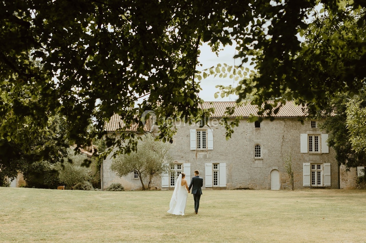 Un couple marche dans un jardin verdoyant en direction d'une grande maison en pierre avec des volets blancs.