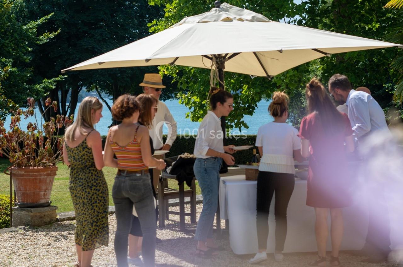 Groupe de personnes discutant près d'un stand sous un parasol dans un jardin ensoleillé.