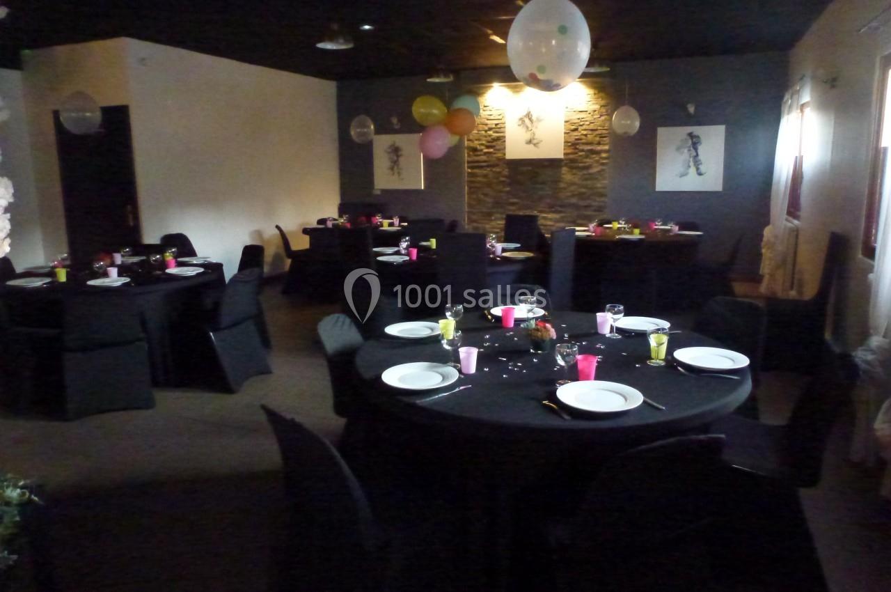 Salle décorée pour un événement, avec des tables rondes dressées, nappes noires et ballons suspendus.