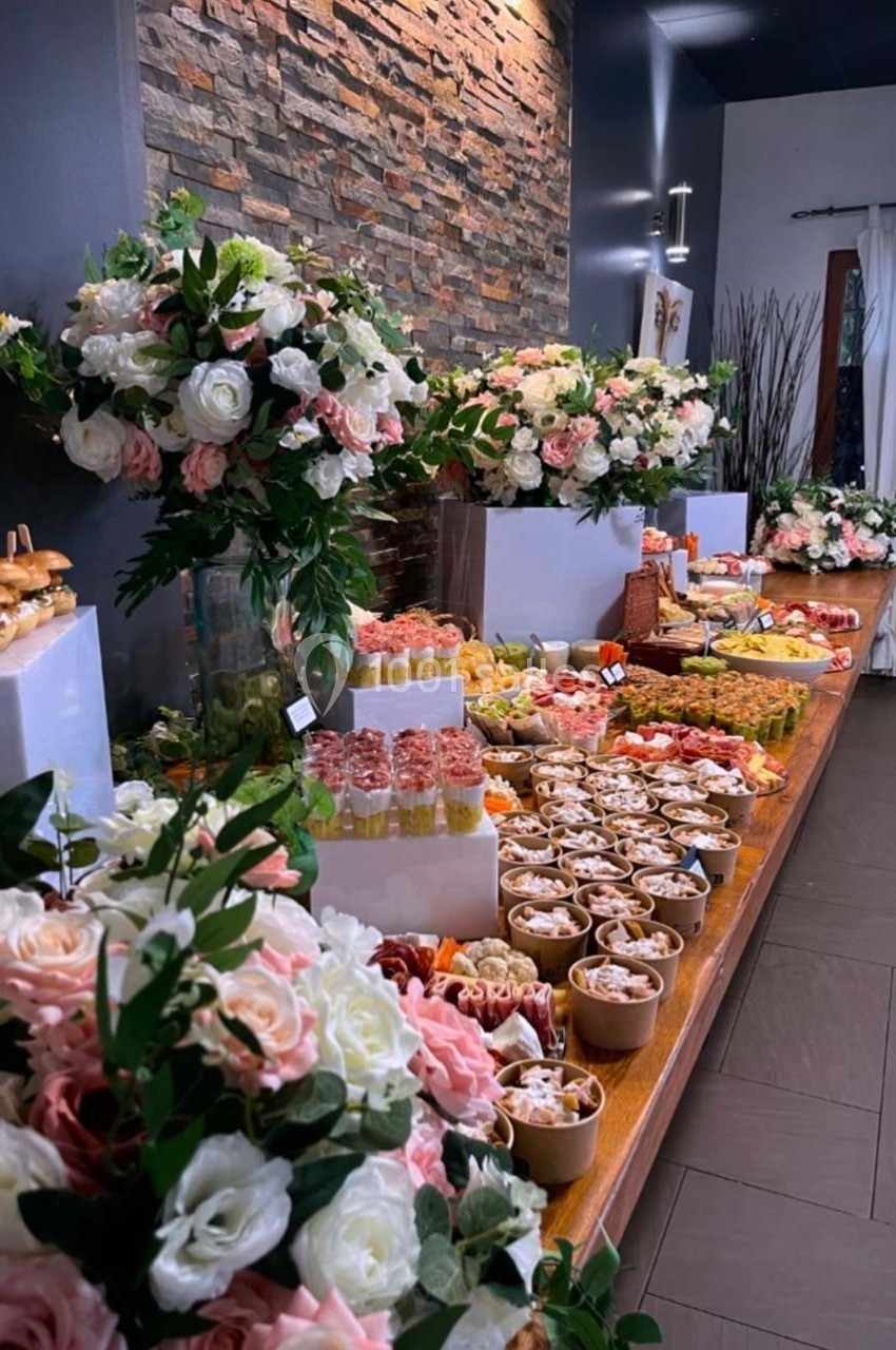 Table garnie de plats variés, desserts et fleurs décoratives, disposée dans une salle au mur de pierre.