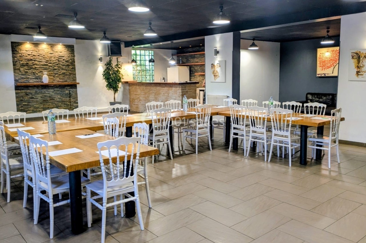 Salle lumineuse avec de grandes tables en bois, chaises blanches, murs décorés et un bar en arrière-plan.