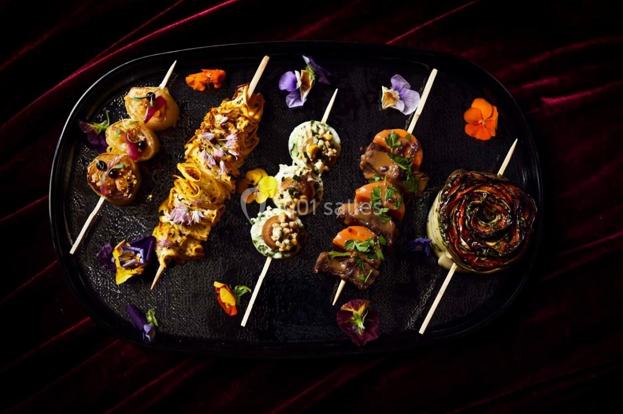 Assortiment de brochettes gastronomiques et roulé de légumes, décorés de fleurs comestibles sur un plateau noir.