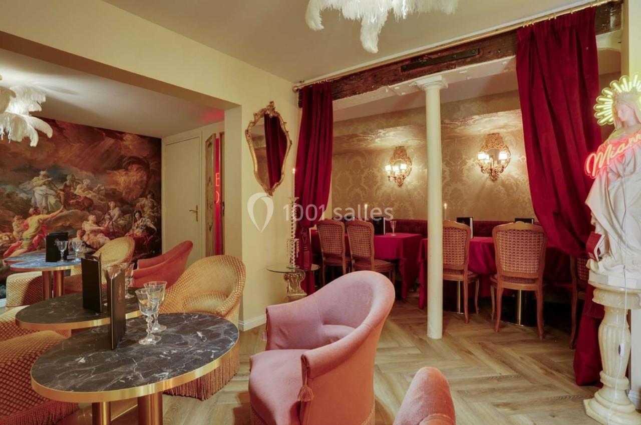 Salon élégant avec fauteuils colorés, tables en marbre, rideaux rouges et fresque murale dans une ambiance chaleureuse.