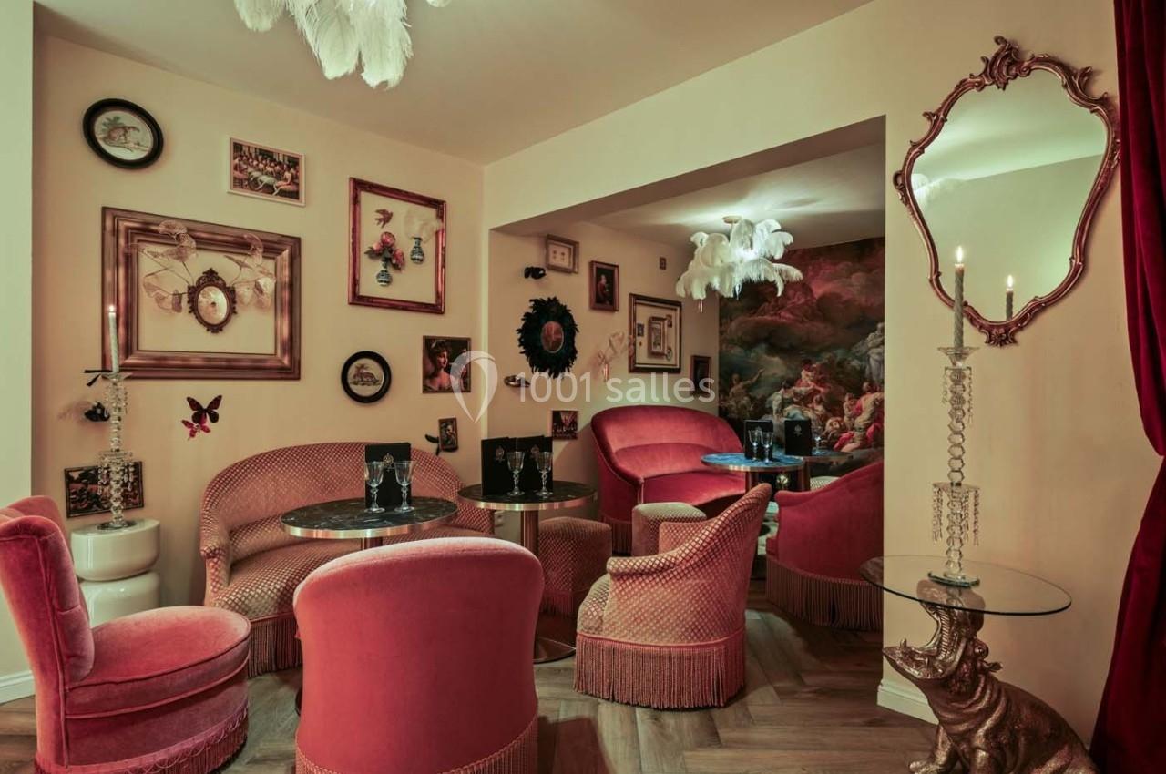 Salon cosy avec fauteuils en velours rose, tables rondes, murs décorés de cadres et éclairage tamisé.