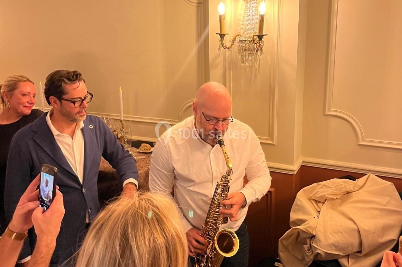 Un homme joue du saxophone dans une pièce éclairée, entouré de plusieurs personnes observant la scène.
