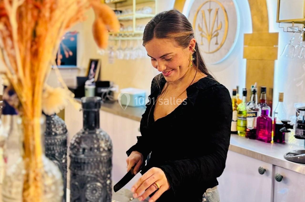 Femme souriante préparant une boisson derrière un comptoir de bar, entourée de bouteilles et de décorations.