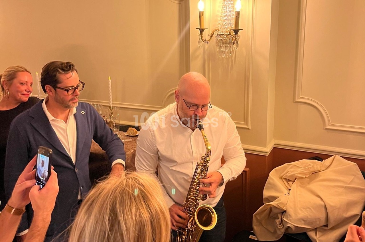 Un homme joue du saxophone dans une pièce élégante, entouré de personnes observant ou prenant des photos.