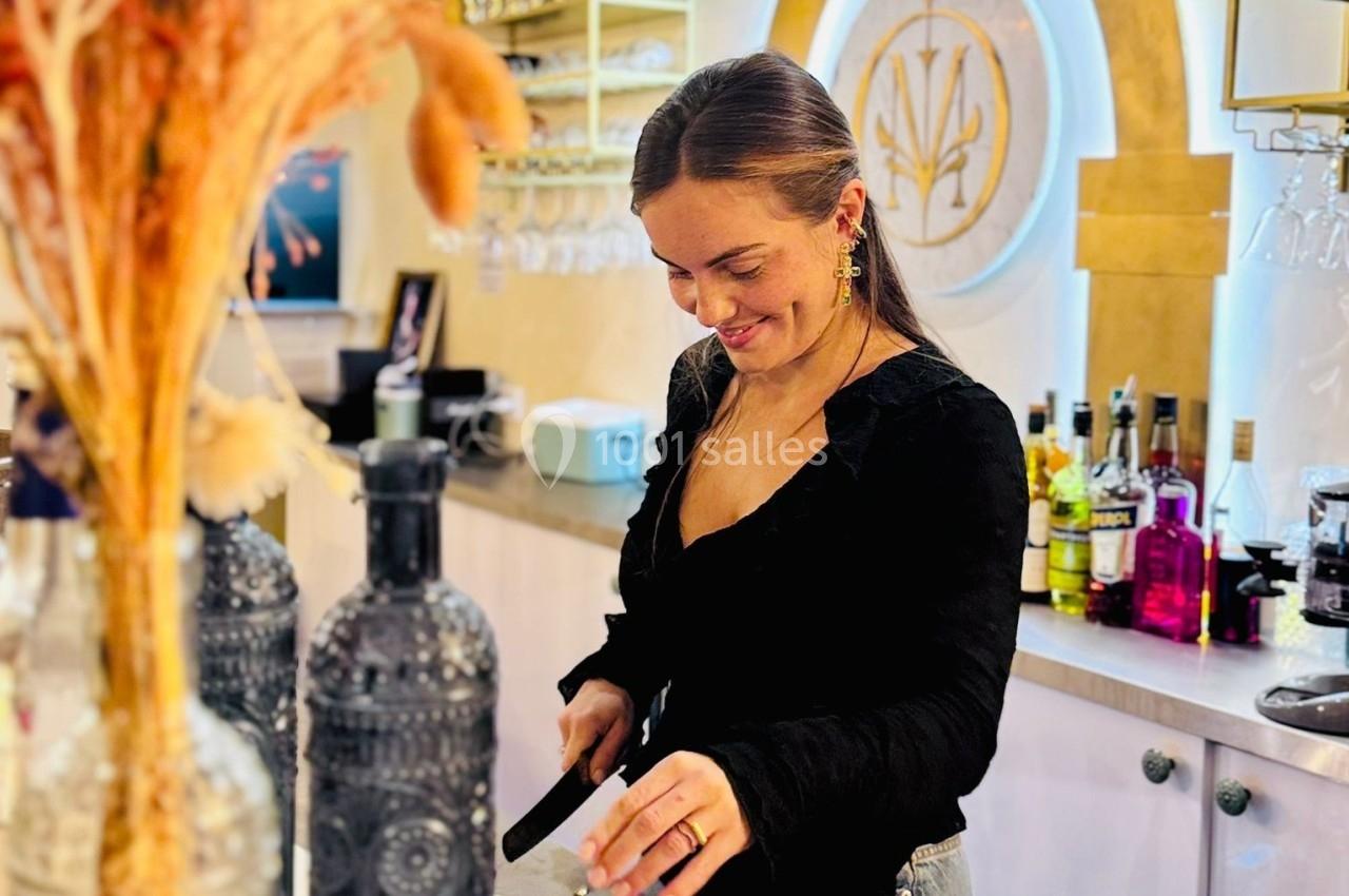 Une femme souriante prépare un cocktail derrière un bar décoré avec des bouteilles et des fleurs séchées.