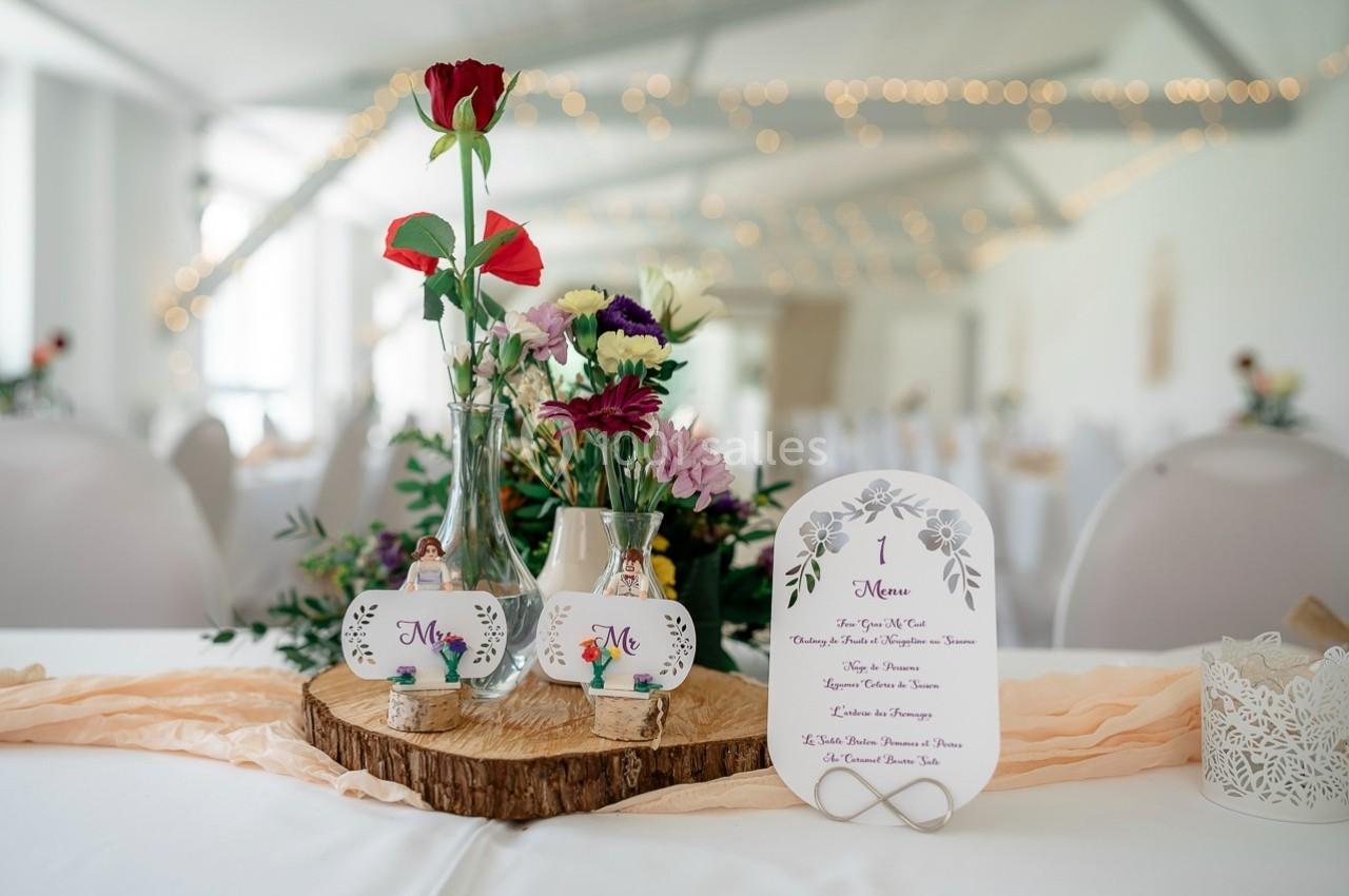 Décoration de table de mariage avec fleurs colorées, menu imprimé et éléments en bois dans une salle lumineuse.
