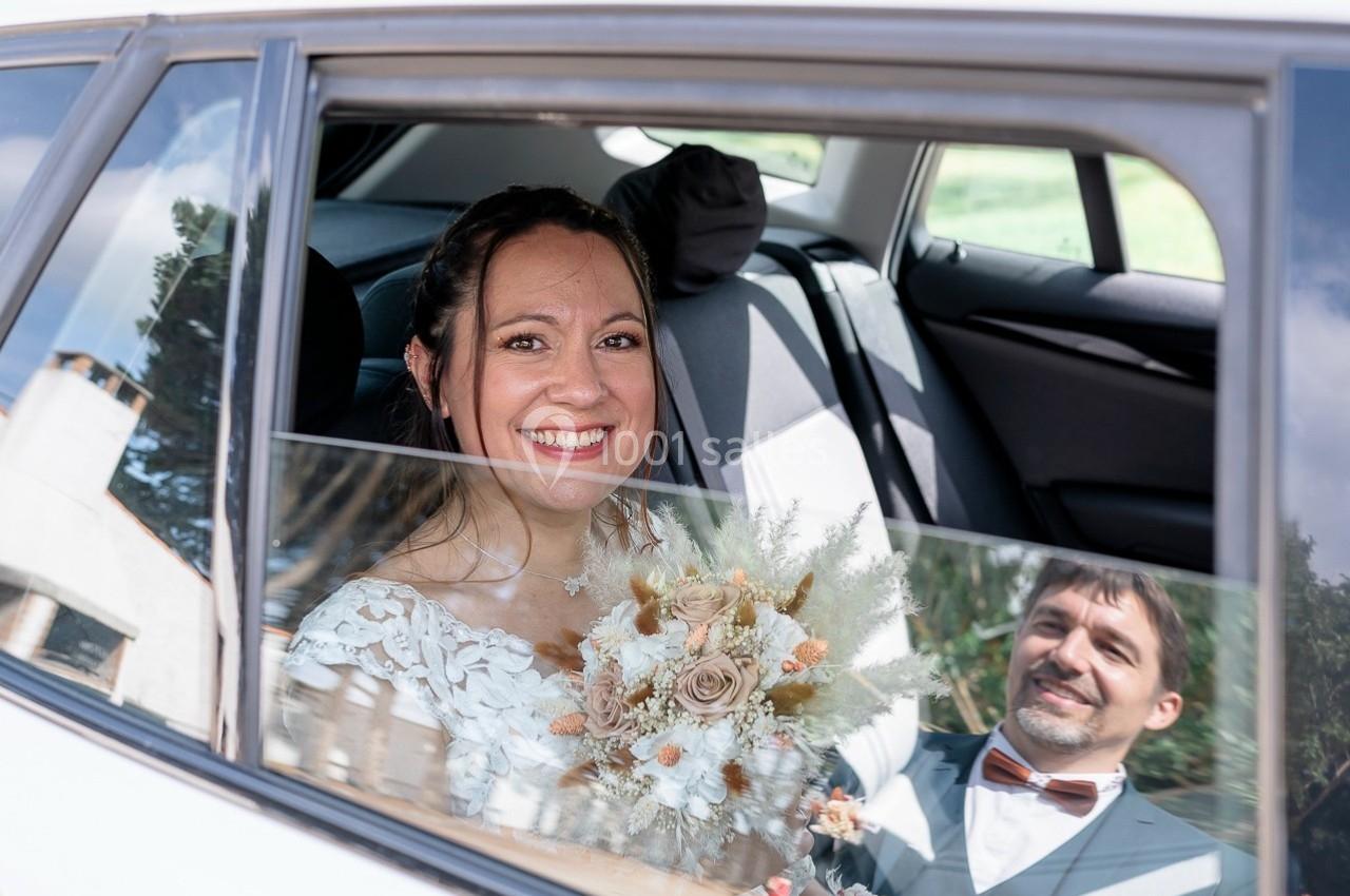 Une mariée souriante tient un bouquet dans une voiture, avec un homme en costume assis à côté d'elle.