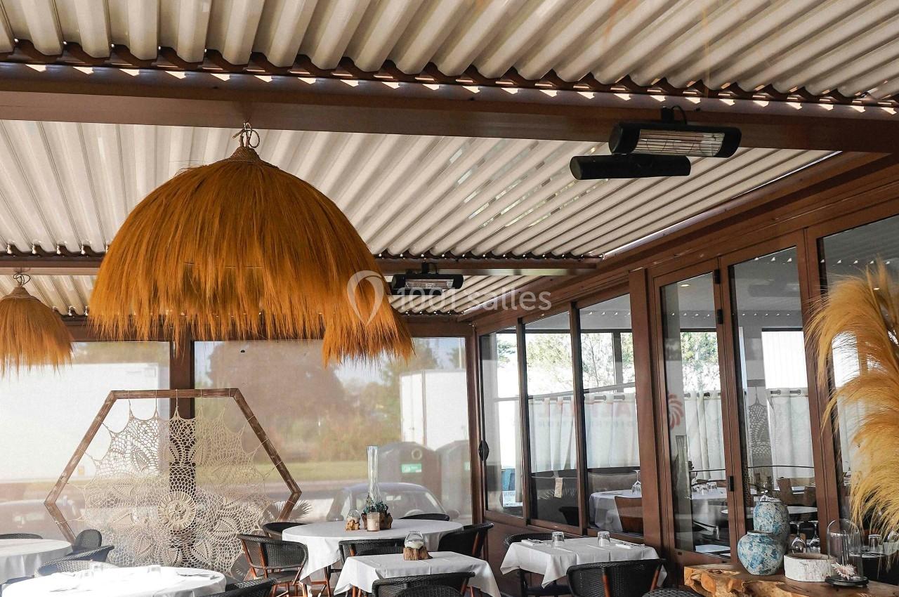 Salle de restaurant lumineuse avec tables dressées, chaises en osier et décorations naturelles sous une pergola.