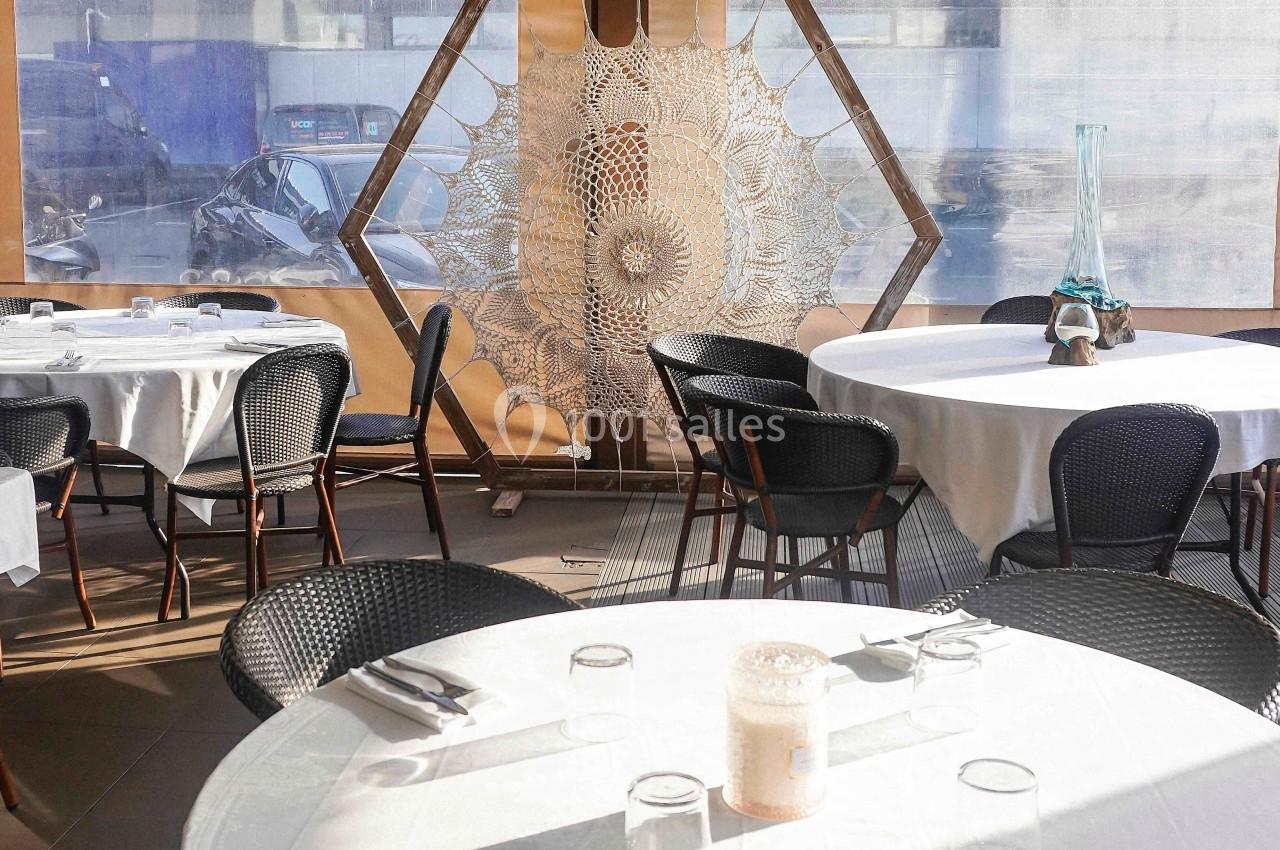 Salle de restaurant lumineuse avec tables rondes dressées, nappes blanches et décoration en macramé sur une fenêtre.