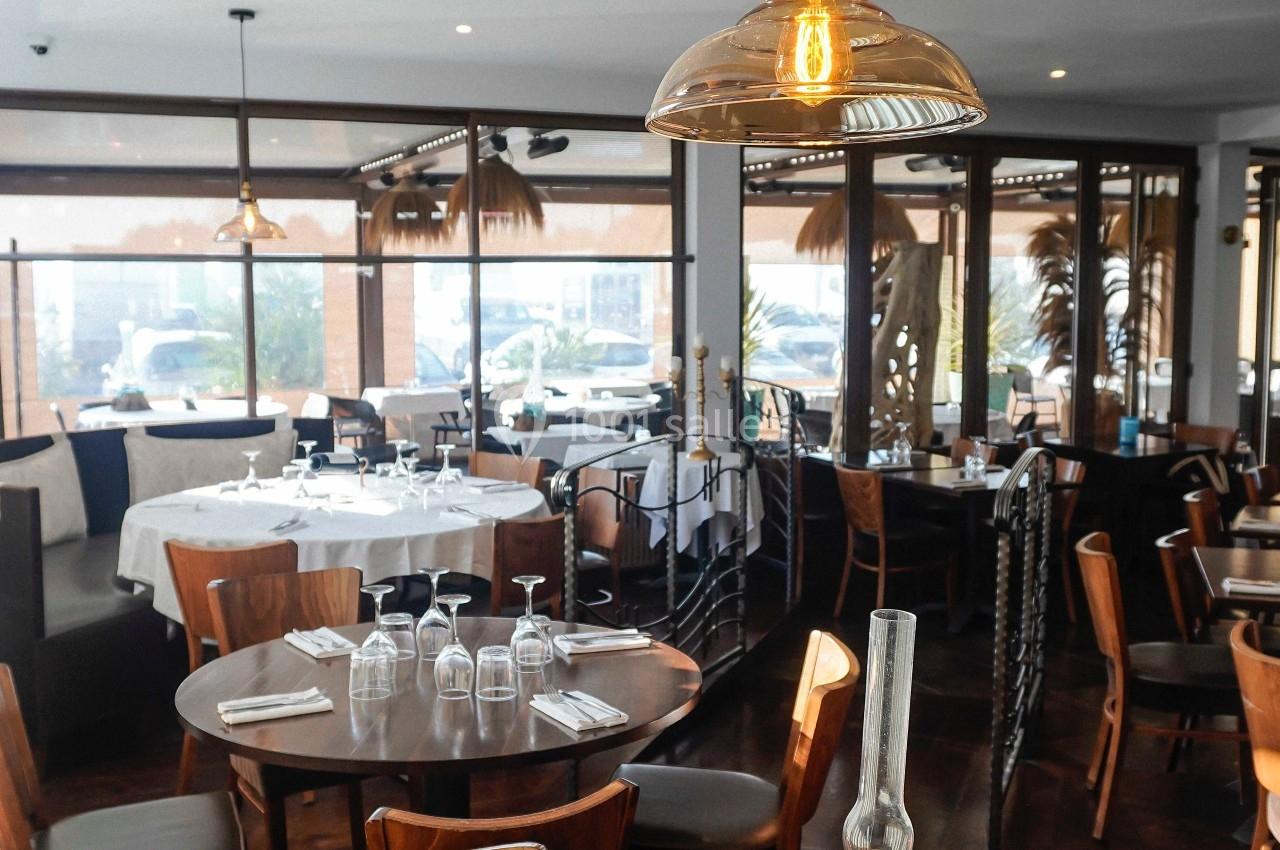 Salle de restaurant lumineuse avec tables en bois dressées, grandes baies vitrées et suspensions en verre.