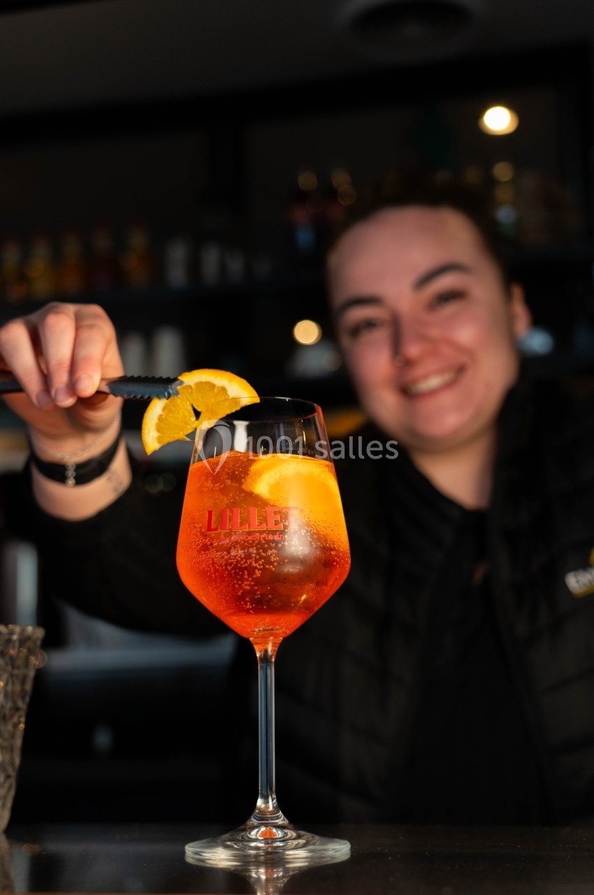 Une personne souriante décore un verre de cocktail orange avec une tranche d'orange dans un bar.