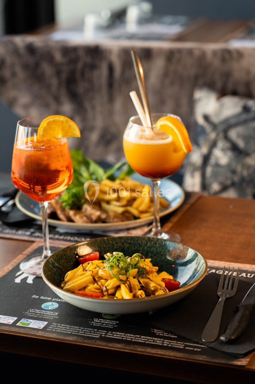 Assiette de pâtes garnie de légumes et herbes fraîches, accompagnée de cocktails et d'un plat avec frites en arrière-plan.
