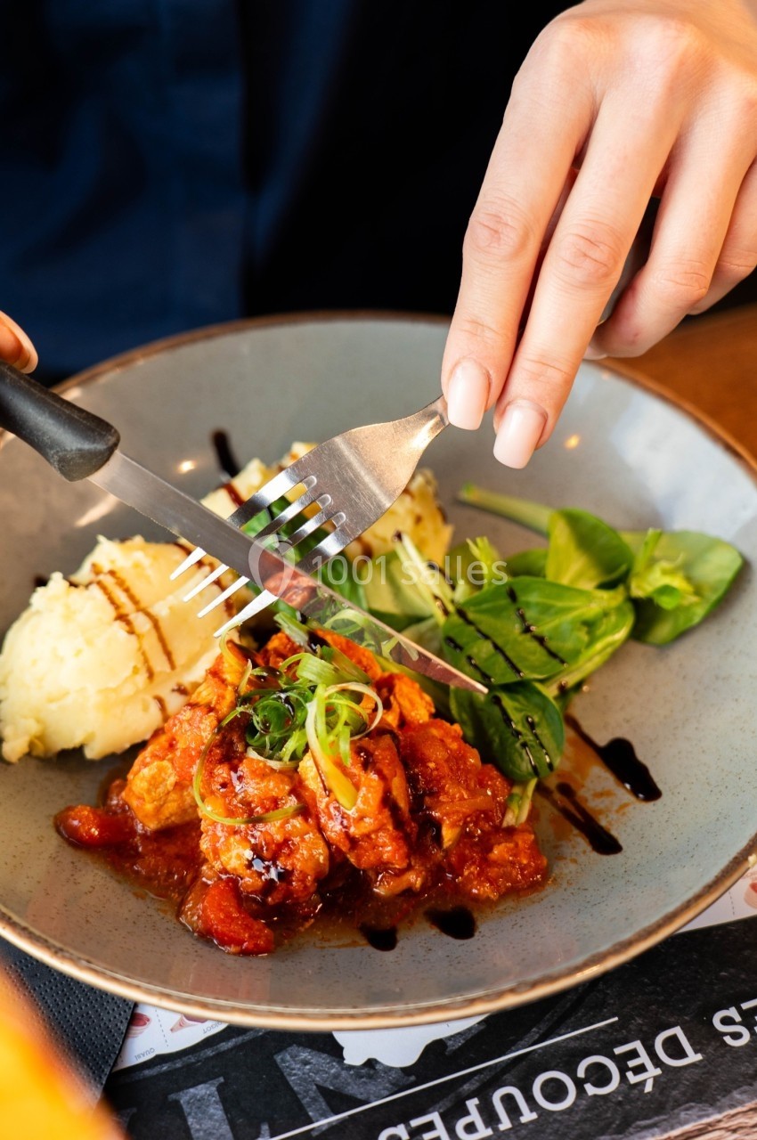 Une assiette contenant un plat de poulet en sauce tomate, purée de pommes de terre et salade verte, avec une main tenant des…