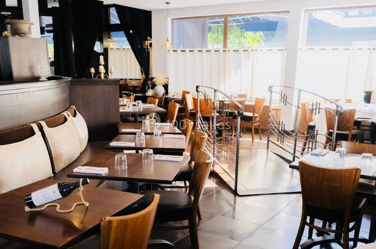 Salle de restaurant lumineuse avec tables en bois, chaises assorties et grande baie vitrée laissant entrer la lumière…