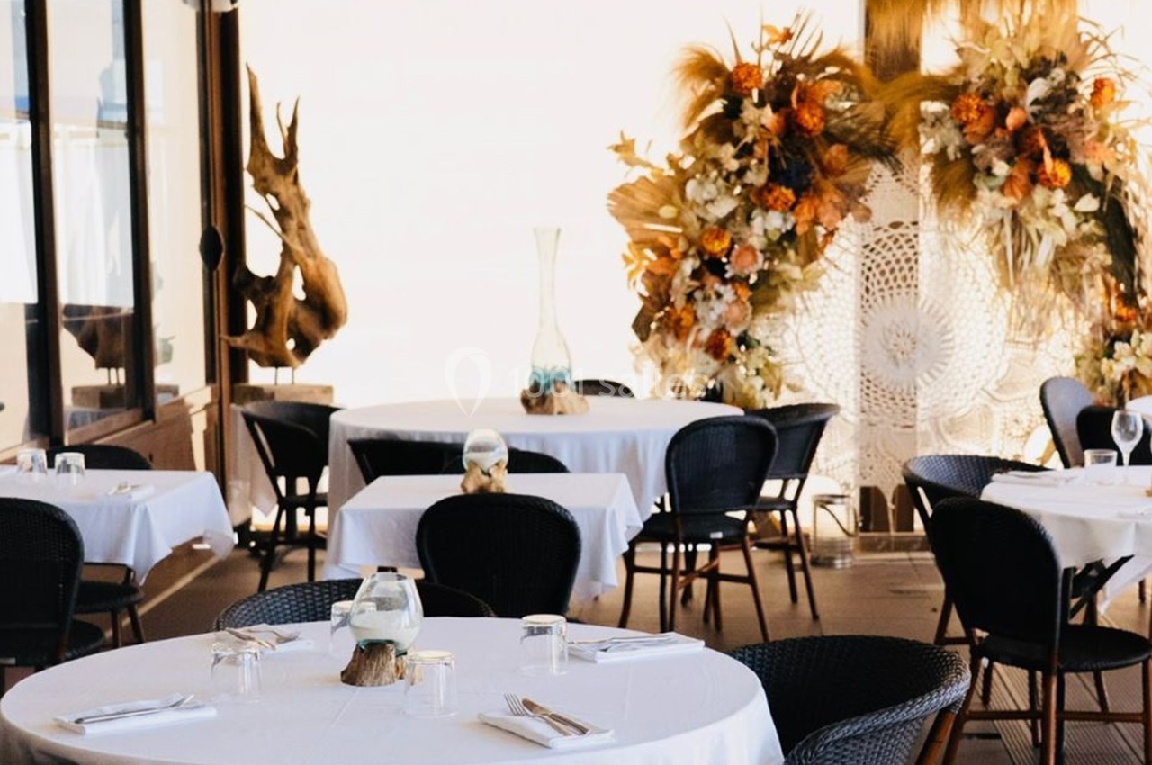 Salle de restaurant décorée avec des tables rondes, nappes blanches et une arche florale en arrière-plan.