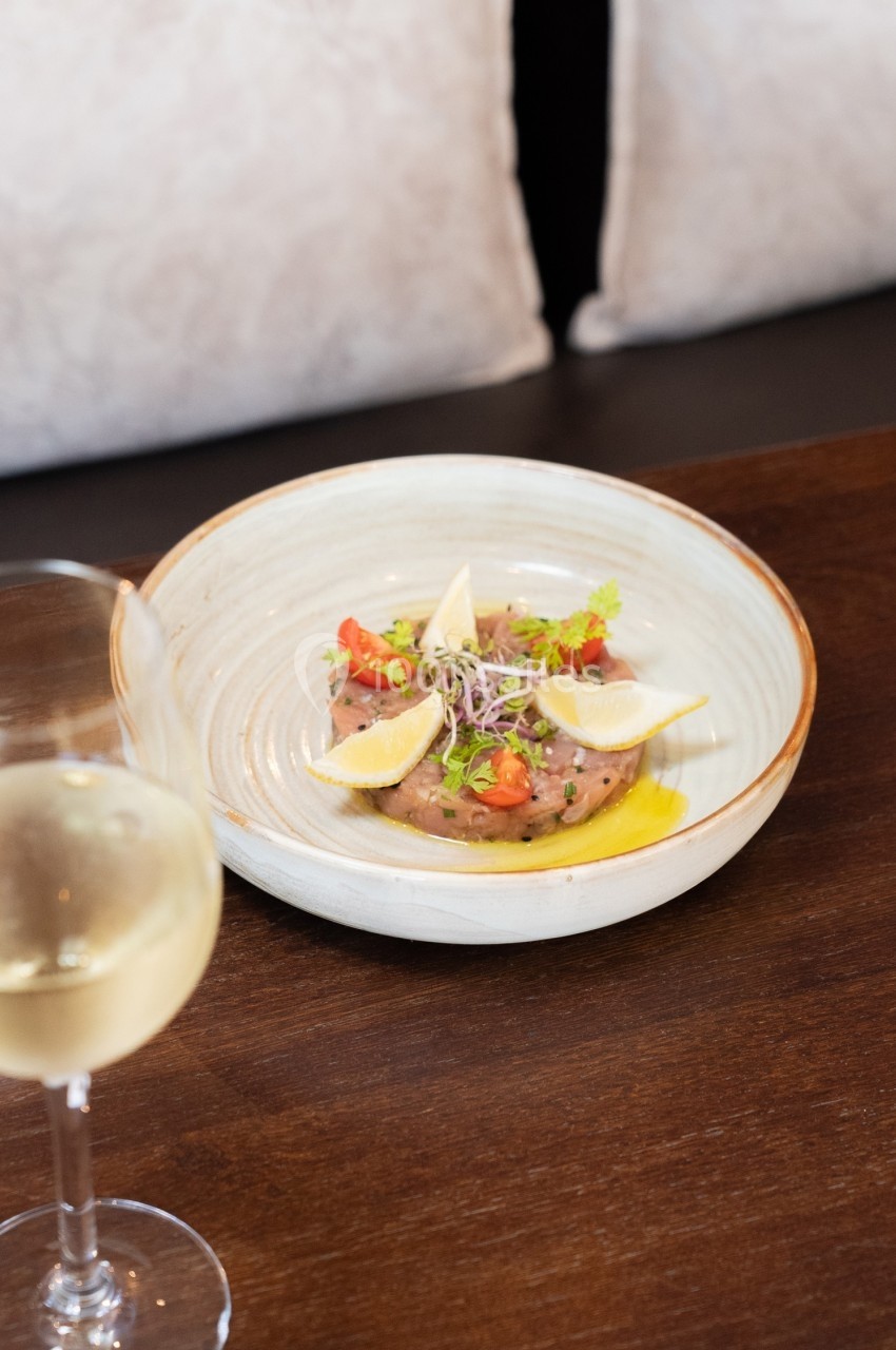 Assiette de tartare de poisson garnie de citron, herbes fraîches et tomates, posée sur une table en bois avec un verre de…