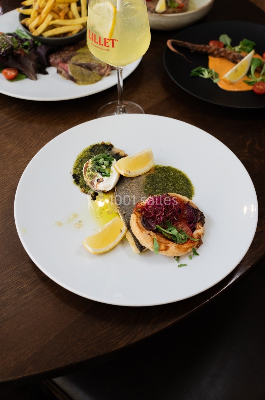 Assiette contenant un poisson grillé, sauce verte, légumes, citron et accompagnements, sur une table en bois.