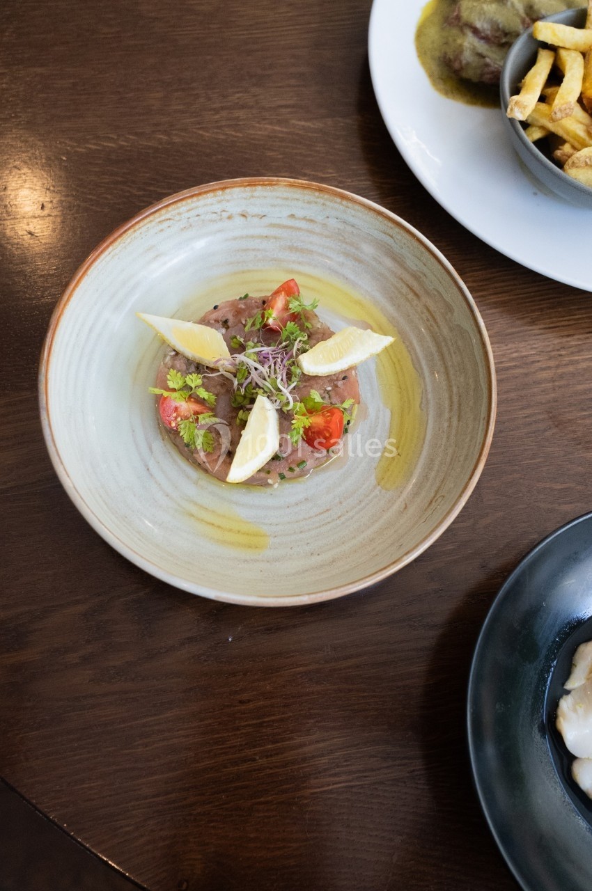 Assiette de tartare de viande garnie de quartiers de citron, tomates cerises et pousses, présentée sur une table en bois.