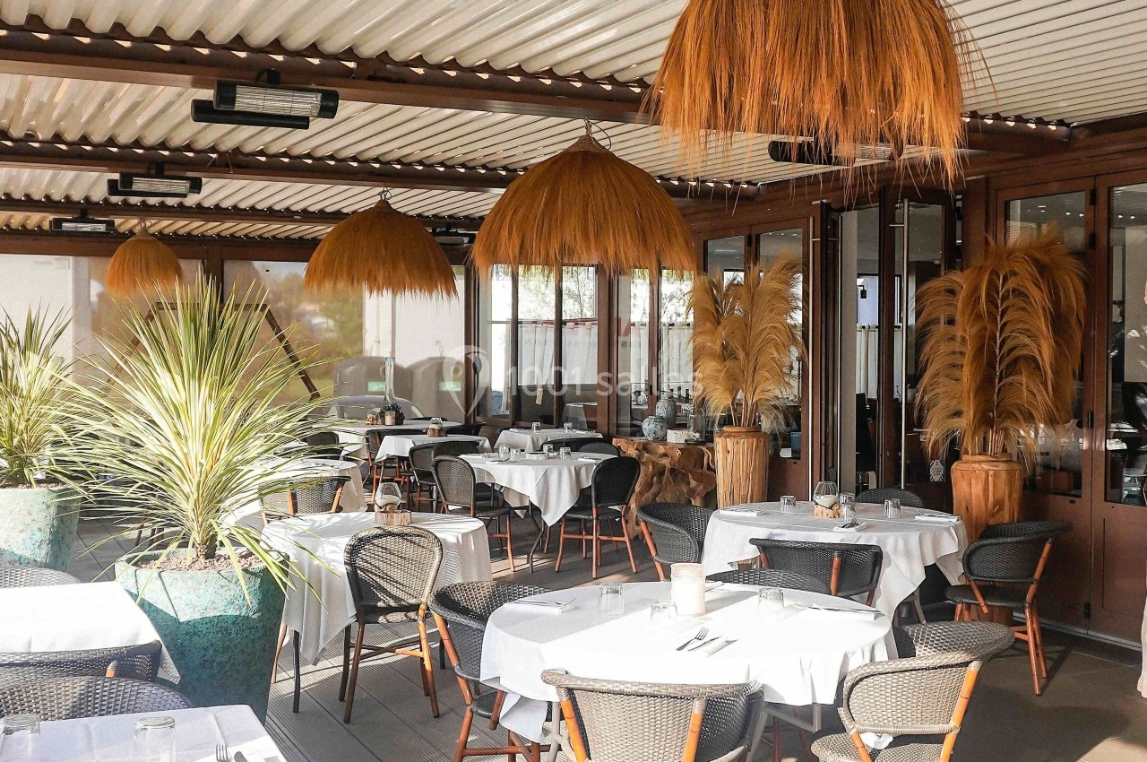 Terrasse de restaurant élégante avec tables dressées, chaises en rotin, plantes décoratives et luminaires suspendus.