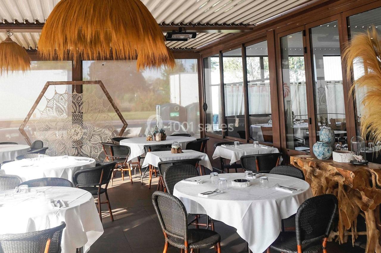 Salle de restaurant lumineuse avec tables rondes dressées, chaises en osier et décoration naturelle en bois et paille.