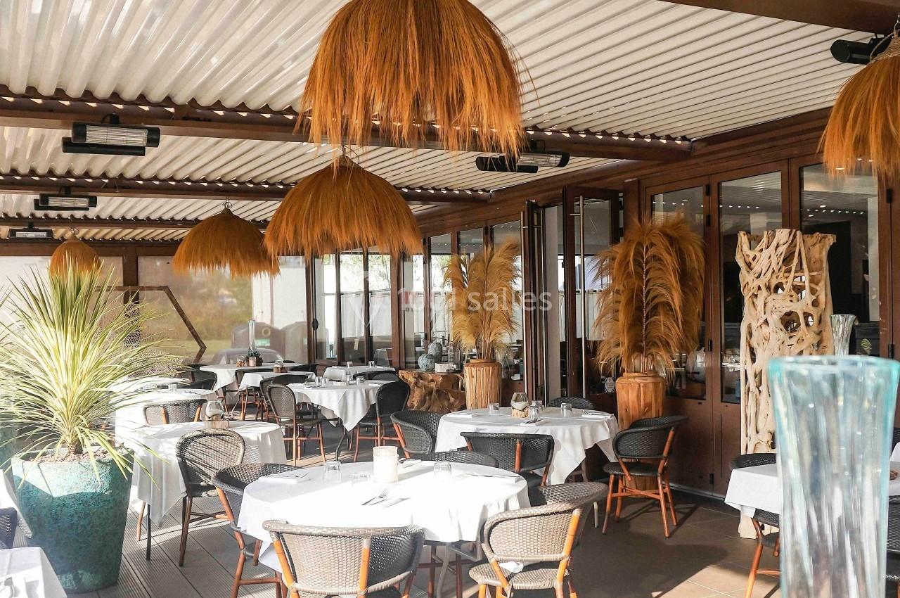 Terrasse de restaurant élégante avec tables dressées, chaises en rotin et décoration végétale sous un toit en pergola.