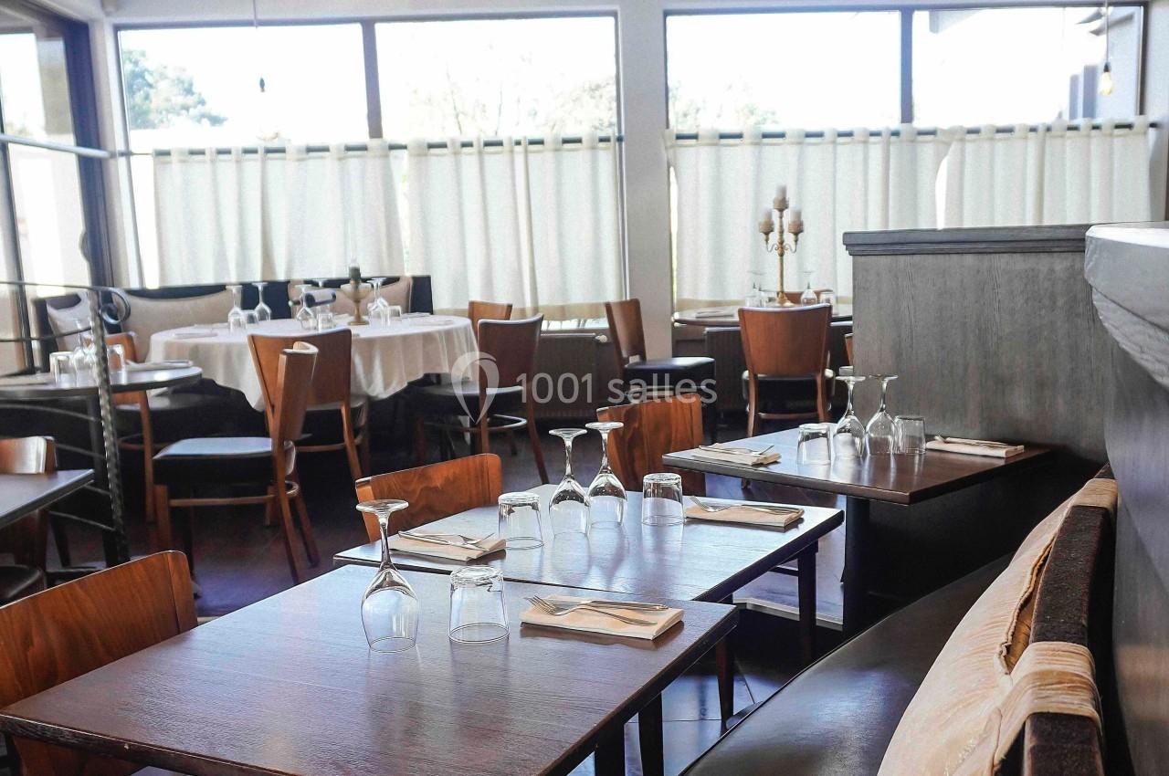 Salle de restaurant lumineuse avec tables en bois dressées, chaises assorties et grandes fenêtres avec rideaux blancs.