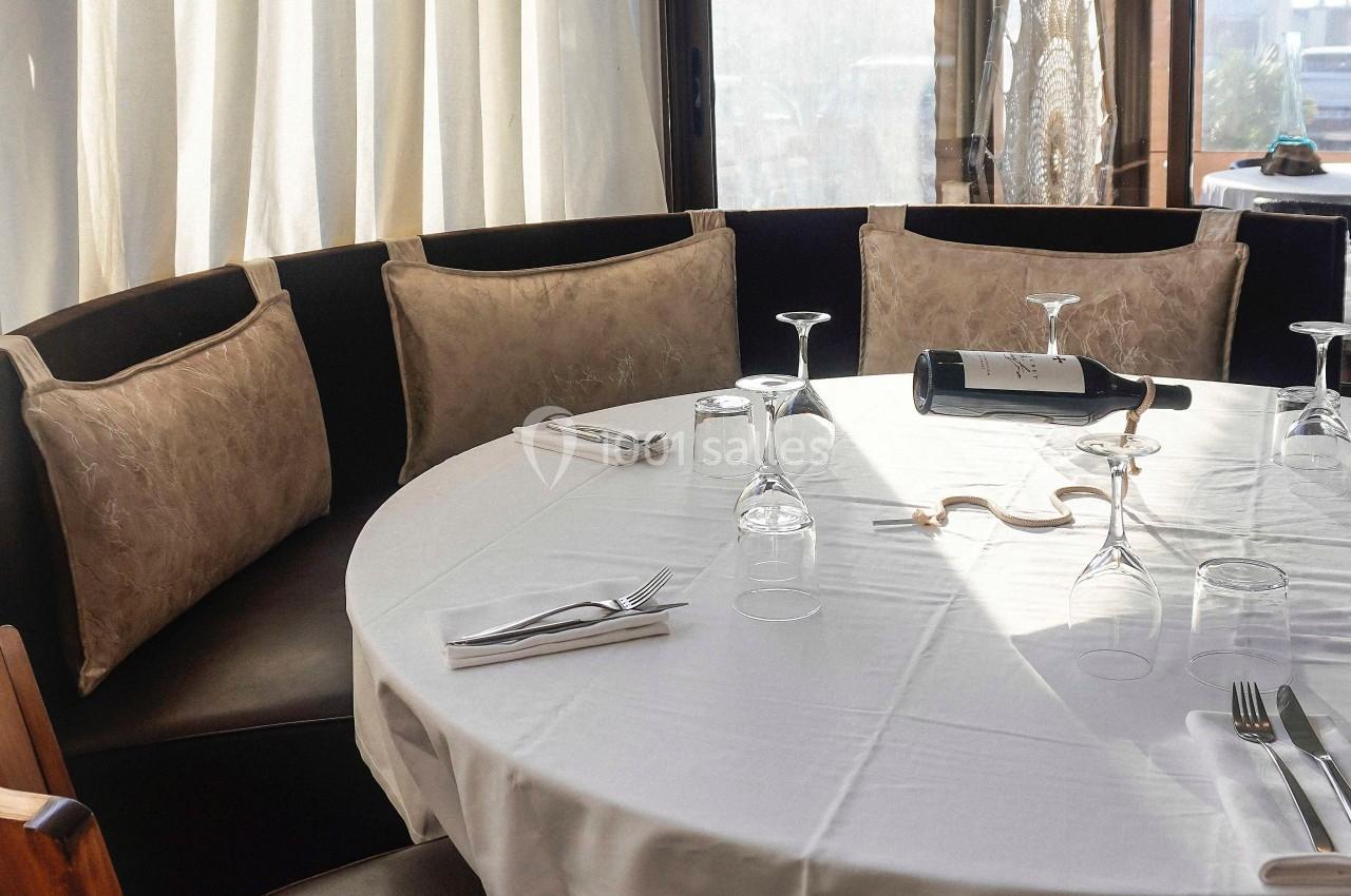 Table ronde dressée avec une nappe blanche, verres, couverts et une bouteille de vin, entourée de banquettes.