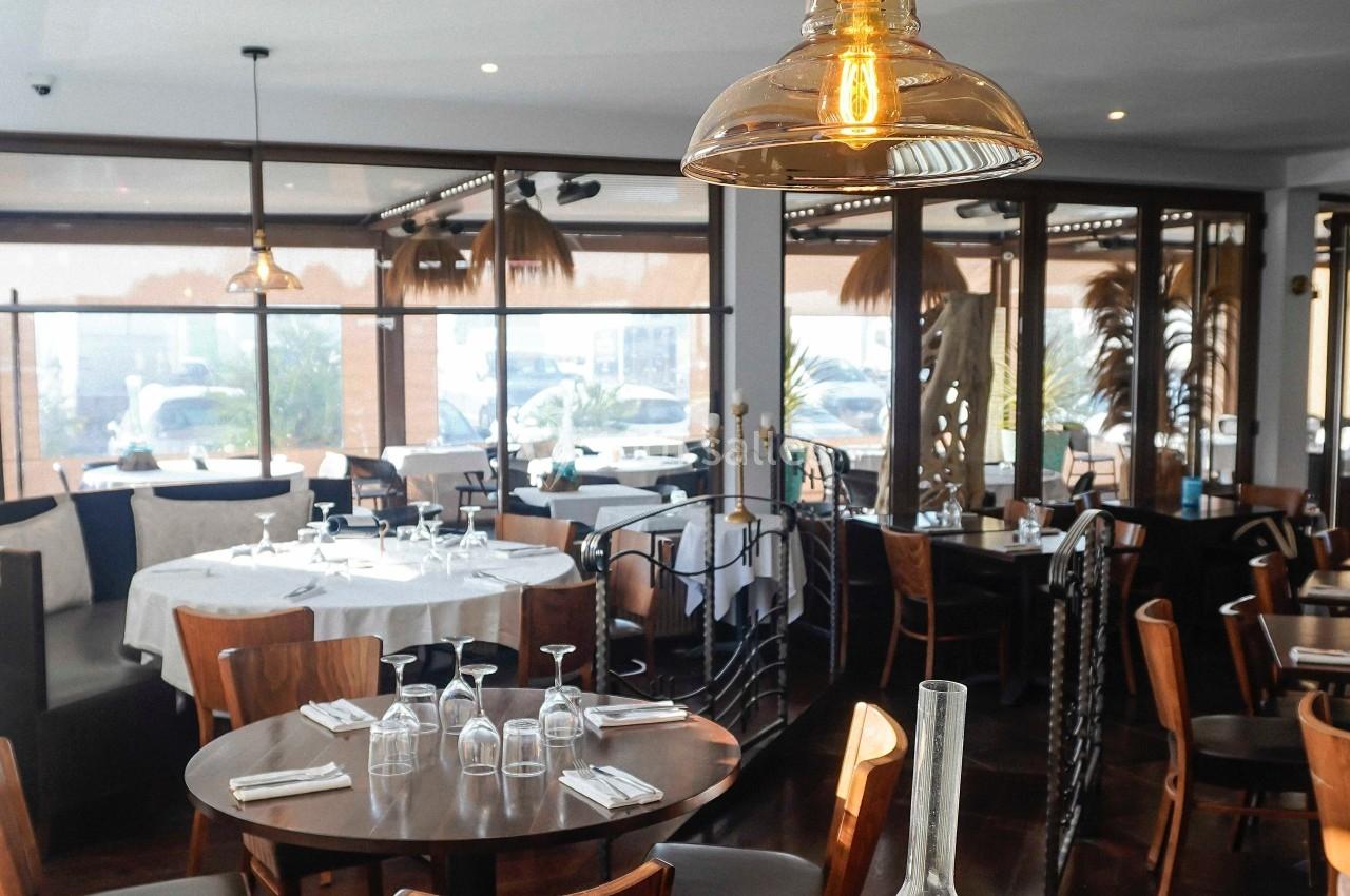 Salle de restaurant lumineuse avec tables dressées, chaises en bois et grandes baies vitrées donnant sur l'extérieur.