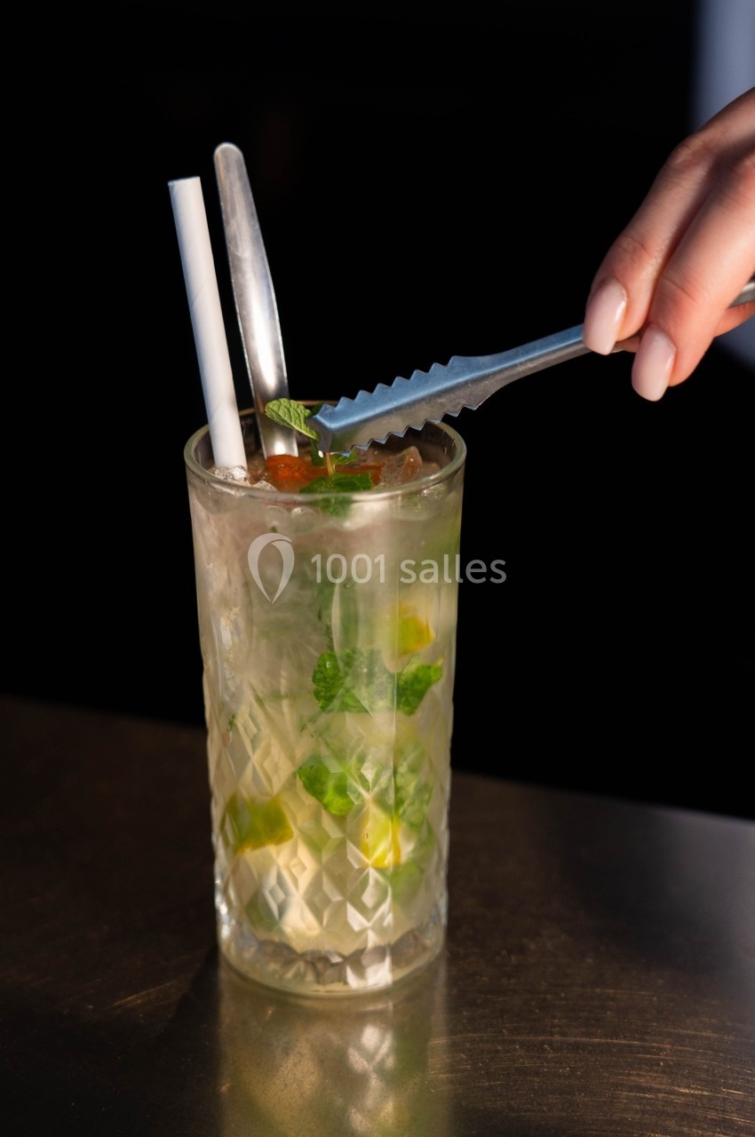 Main ajoutant une feuille de menthe à un cocktail glacé avec des pailles, dans un verre transparent à motifs.