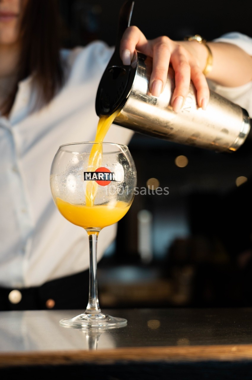 Une personne verse un cocktail orange dans un verre à pied à l'aide d'un shaker en métal.