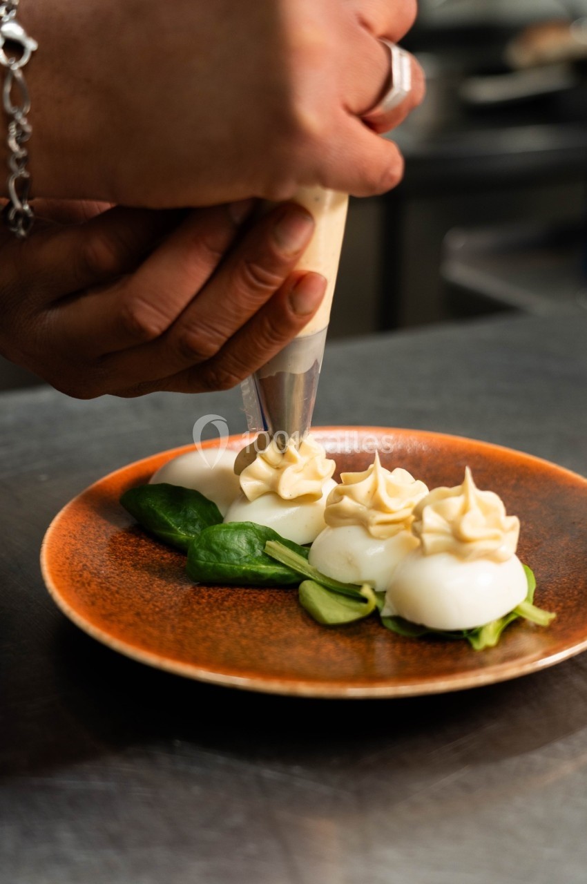Une main décore des œufs pochés avec une poche à douille sur une assiette garnie de feuilles vertes.