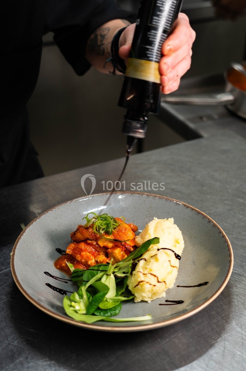 Un chef ajoute une touche de sauce balsamique sur une assiette contenant de la purée, de la viande en sauce et de la salade.
