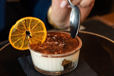 Dessert tiramisu dans une verrine, décoré d'une tranche d'orange séchée, avec une main tenant une cuillère.