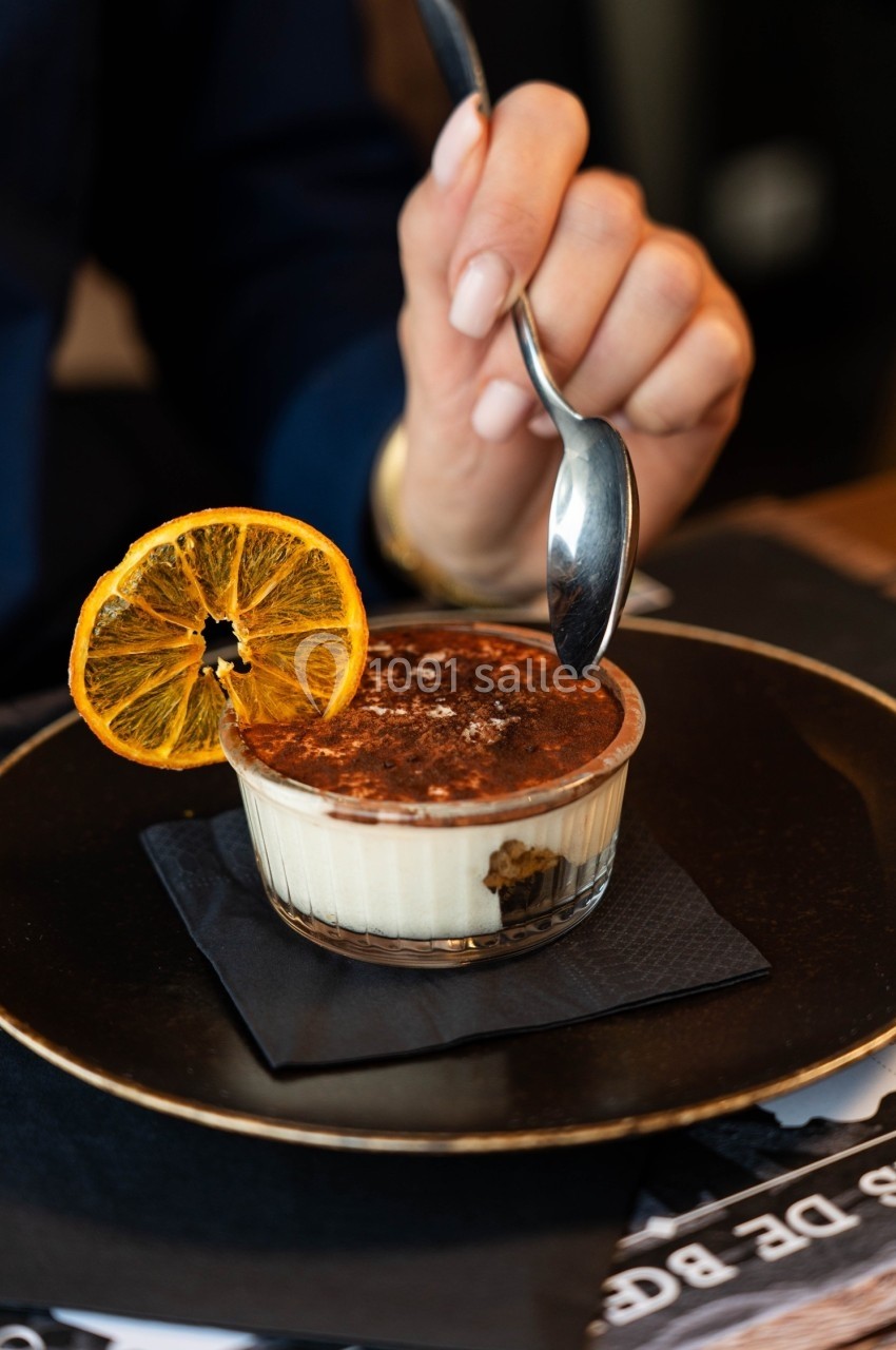 Dessert tiramisu dans une verrine, décoré d'une tranche d'orange séchée, avec une main tenant une cuillère.