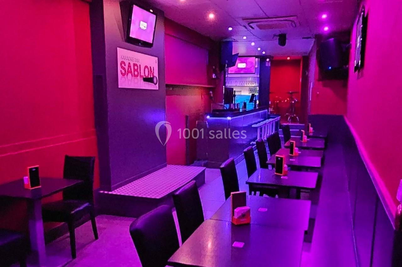 Salle de bar avec tables alignées, éclairage violet et rouge, écrans muraux et comptoir au fond.