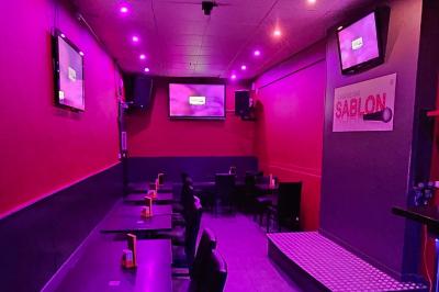 Écran et panneau indiquant ’Karaoké Bar Sablon’ avec un micro, dans une salle éclairée par des lumières violettes.