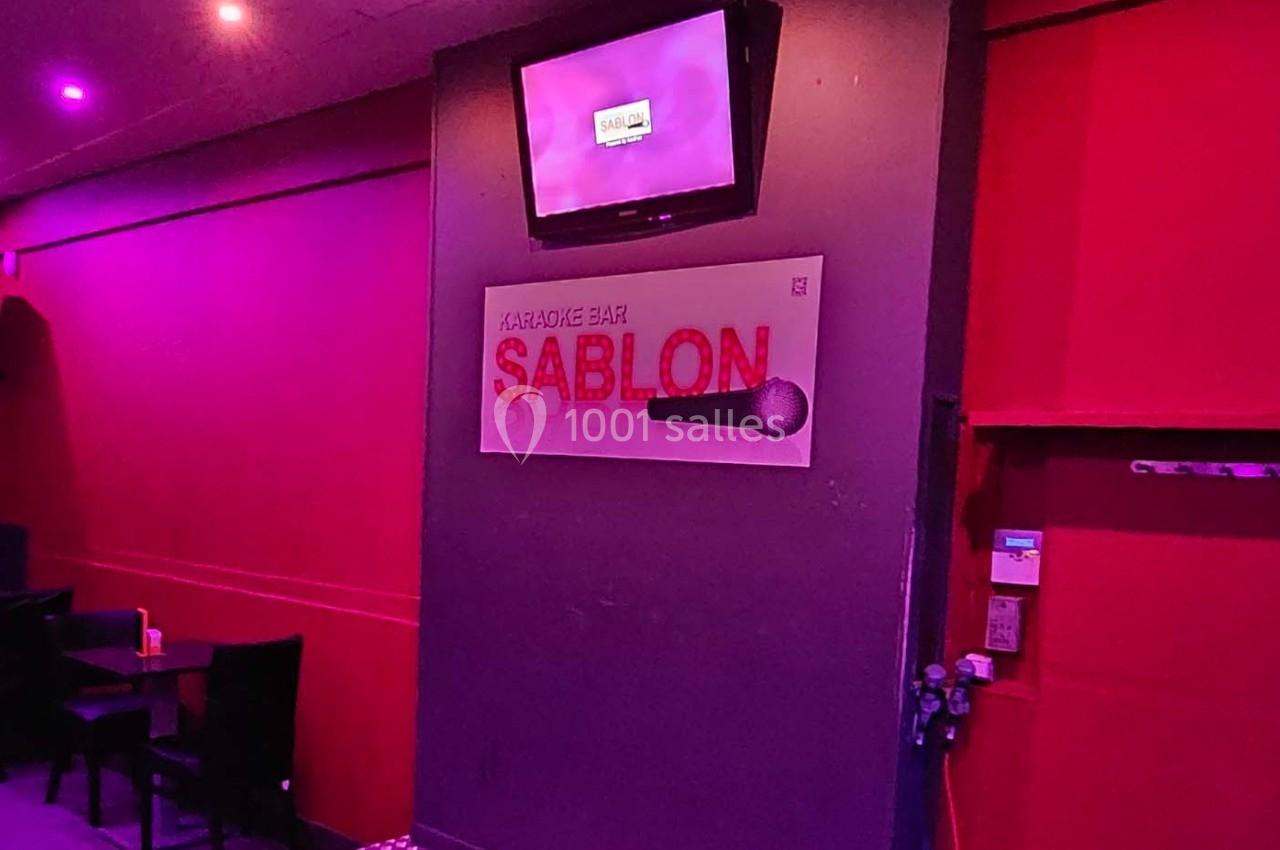 Écran et panneau indiquant ’Karaoké Bar Sablon’ avec un micro, dans une salle éclairée par des lumières violettes.