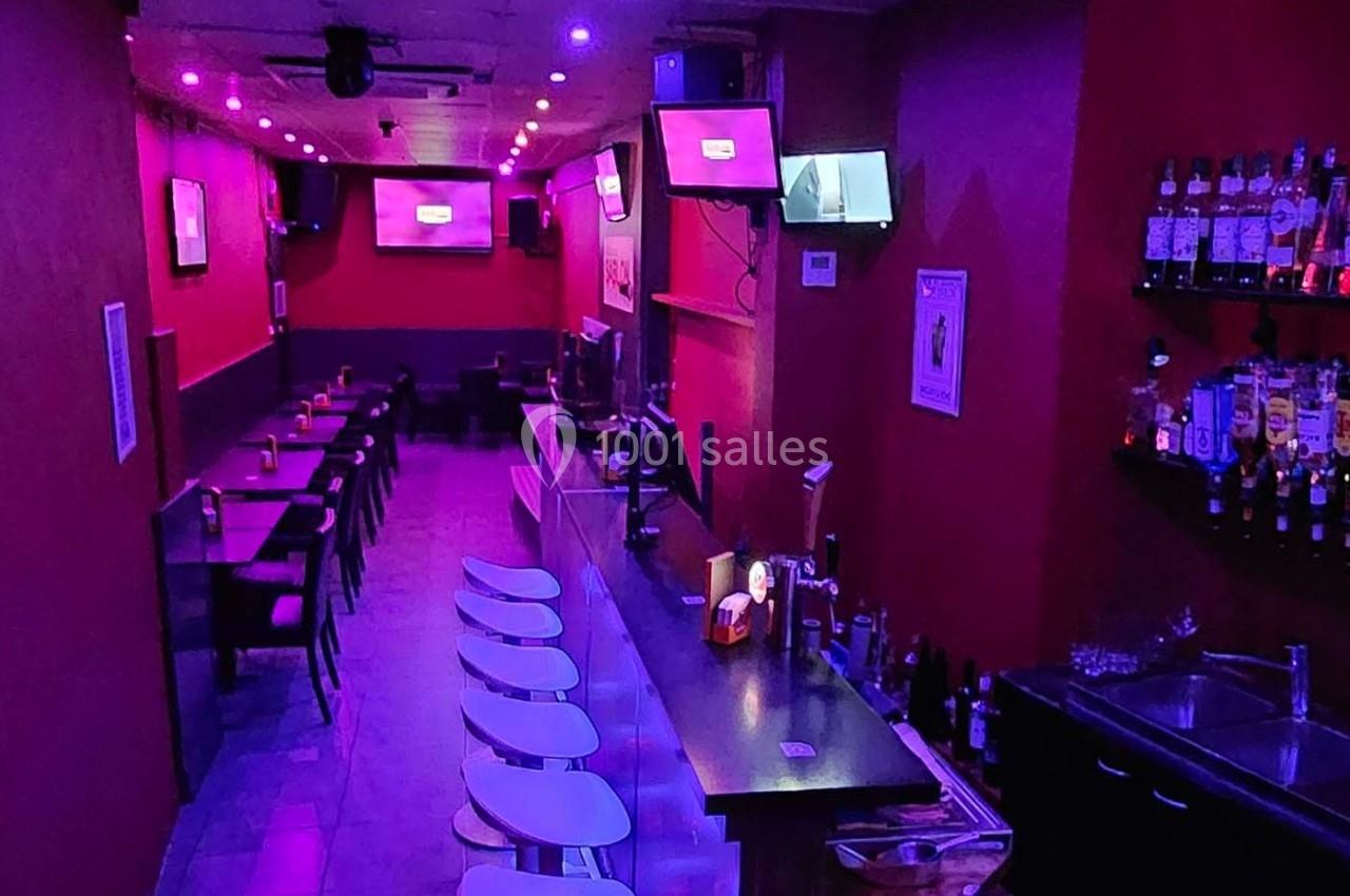 Bar à l'éclairage violet avec comptoir, tabourets, tables alignées et étagères de bouteilles sur un mur rouge.