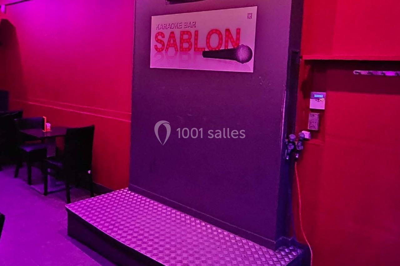 Scène de karaoké dans un bar avec éclairage rose et rouge, micro illustré sur un panneau mural.