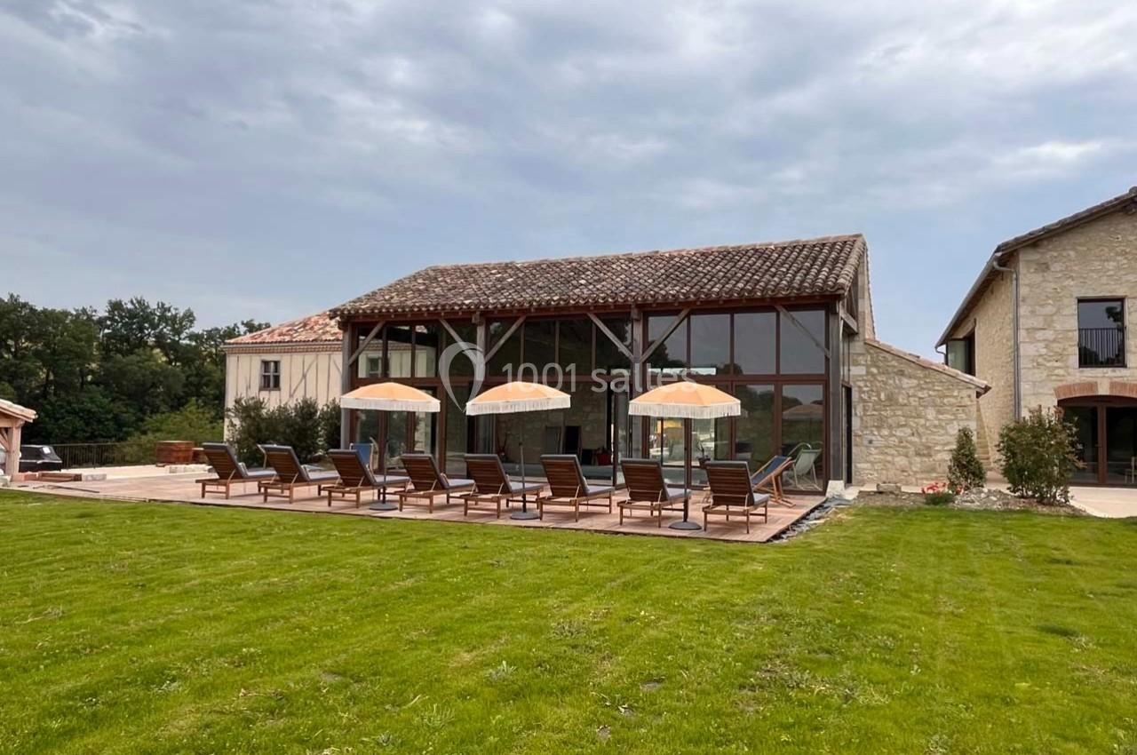 Grande maison en pierre avec terrasse aménagée, chaises longues et parasols, entourée de pelouse et végétation.