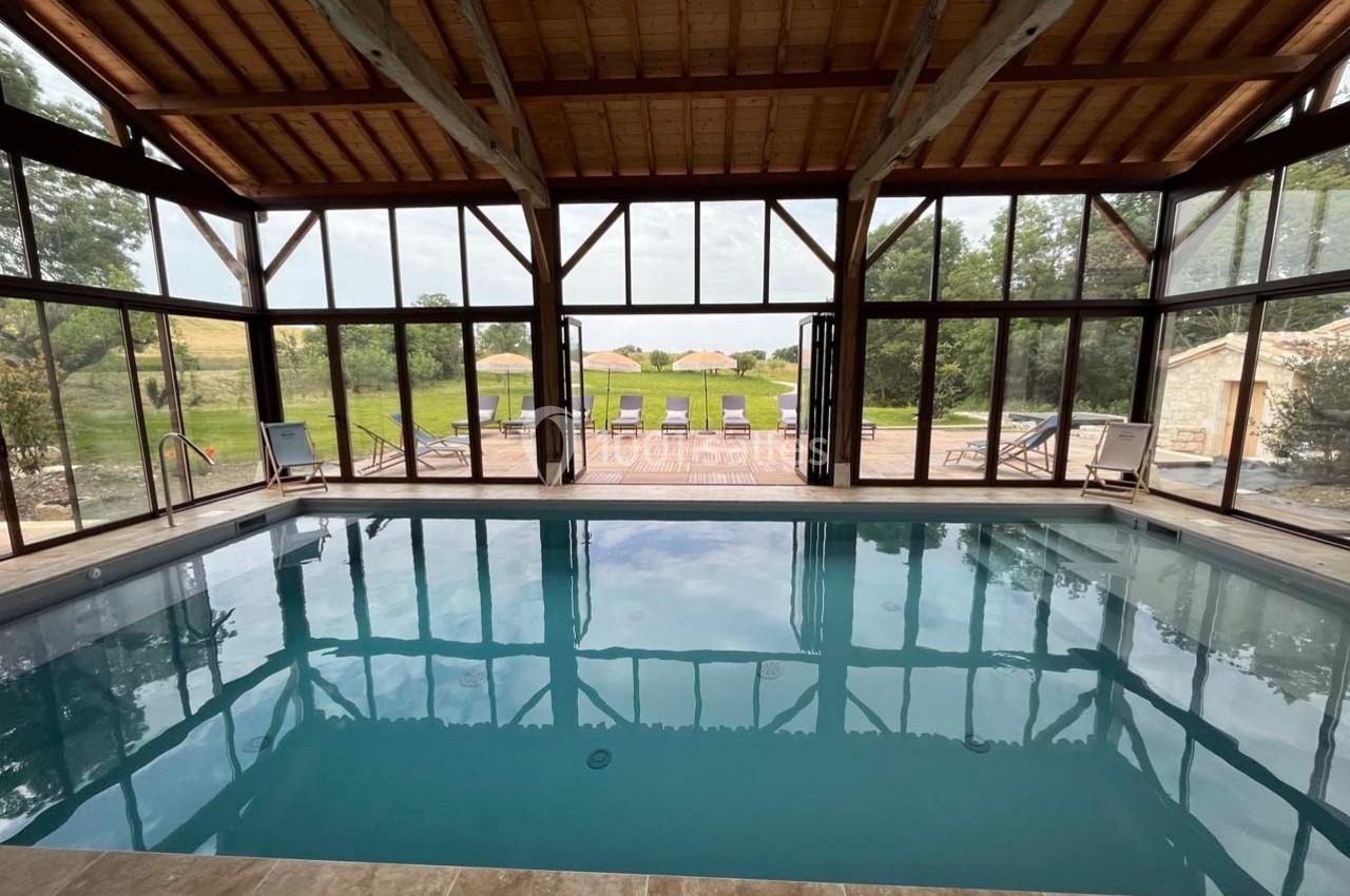 Piscine intérieure avec baies vitrées donnant sur une terrasse en bois, des chaises longues et un paysage verdoyant.