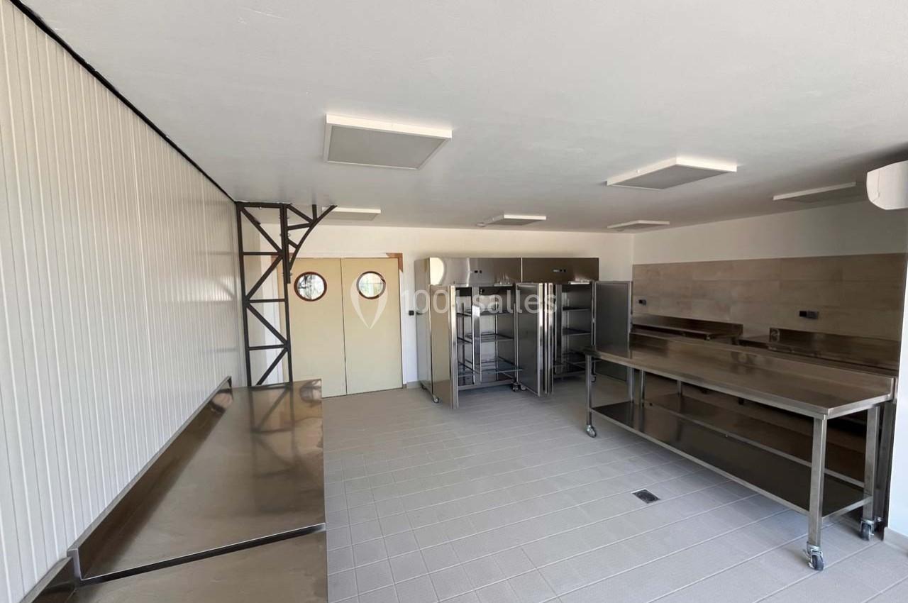 Cuisine professionnelle vide avec plans de travail en inox, étagères métalliques et carrelage au sol.