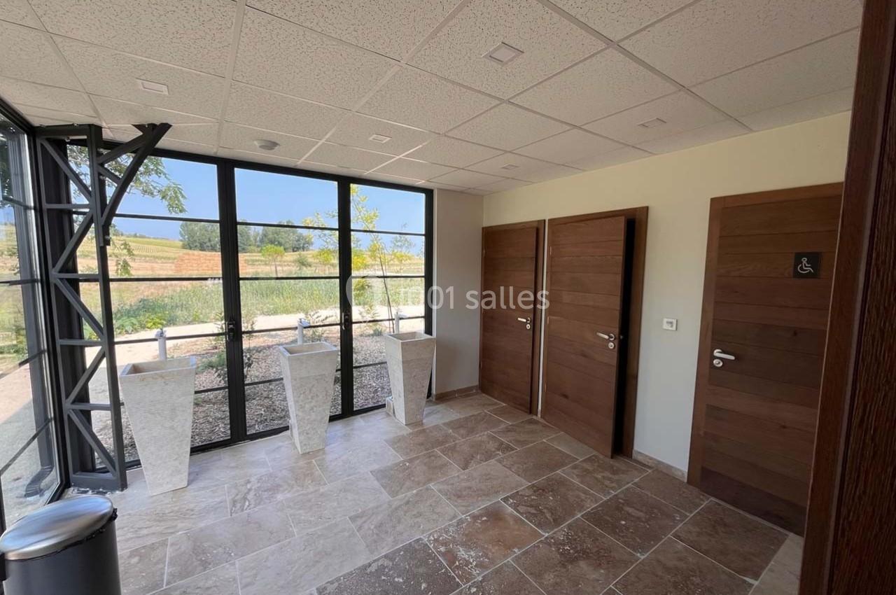 Espace intérieur lumineux avec sol en pierre, trois portes en bois et grandes fenêtres donnant sur un paysage extérieur.