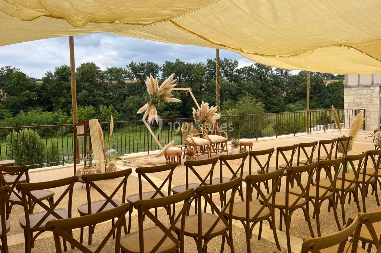 Chaises en bois alignées sous une toile beige, face à une décoration florale et un paysage verdoyant.