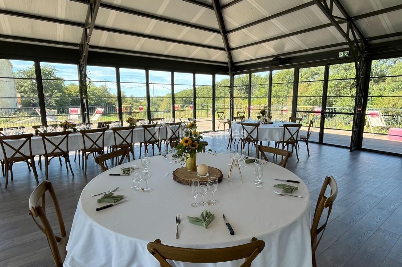 Salle lumineuse avec de grandes baies vitrées, tables rondes dressées avec nappes blanches et décorations florales.
