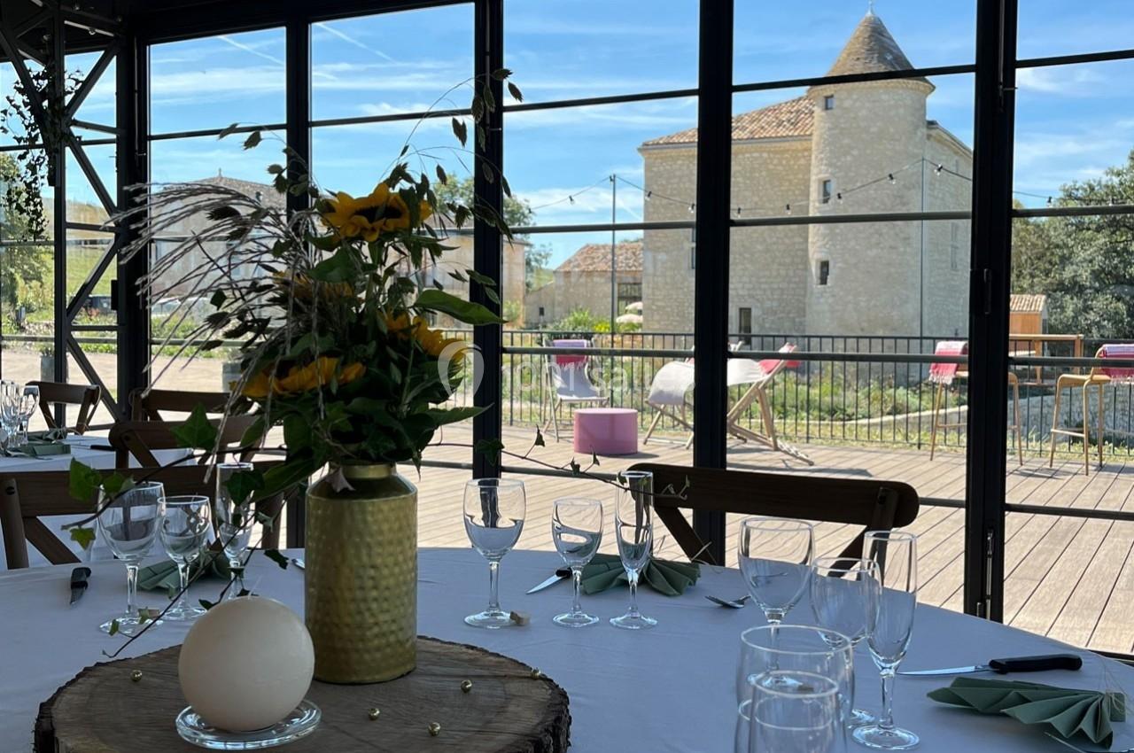 Table dressée avec décoration florale, vue sur une terrasse ensoleillée et un bâtiment en pierre avec une tour.